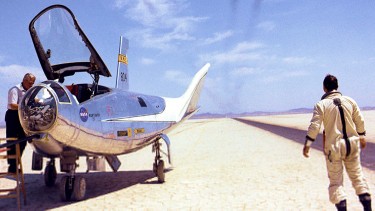 alt="A NASA pilot looks up at a passing aircraft from the sand"