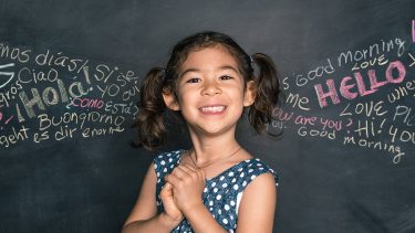 Young girl standing in front of a blackboard with multiple languages written on it.